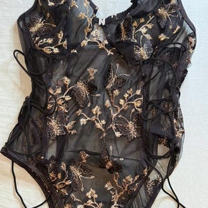 May include: Black lingerie bodysuit with gold floral embroidery and butterfly accents. The sheer fabric features a lace-up design on the sides and adjustable straps. The bodysuit has a delicate, feminine aesthetic.