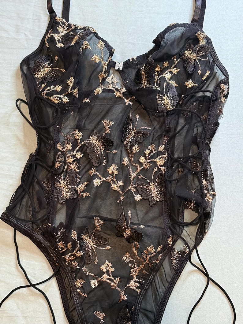 May include: Black lingerie bodysuit with gold floral embroidery and butterfly accents. The sheer fabric features a lace-up design on the sides and adjustable straps. The bodysuit has a delicate, feminine aesthetic.