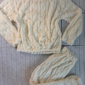 May include: Cream-colored, plush pajama set. The set includes a long-sleeved top and matching pants. The fabric has a textured, cable-knit design. The top has a crew neck and elastic waist. The pants are also elastic waist. The set is made of a soft, fuzzy material.