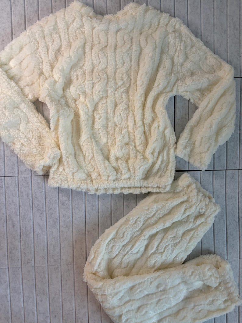 May include: Cream-colored, plush pajama set. The set includes a long-sleeved top and matching pants. The fabric has a textured, cable-knit design. The top has a crew neck and elastic waist. The pants are also elastic waist. The set is made of a soft, fuzzy material.