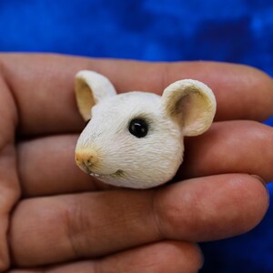 K&ouml;nnte beinhalten: Detaillierte, wei&szlig;e M&auml;usefigur mit strukturierter Oberfl&auml;che, kleinen Ohren und schwarzen Augen. Die Maus hat eine hellrosa Nase. Die Figur wird in einer Hand vor blauem Hintergrund gehalten. Miniaturskulptur.