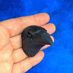 May include: A detailed, black raven head sculpture with textured feathers and a sharp beak. The raven's eye is visible, and the sculpture is held in a hand against a blue background. This piece is likely a decorative item or part of a larger art project.