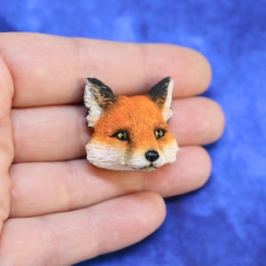 Peut inclure: Une figurine miniature détaillée en forme de tête de renard. Le renard a une fourrure orange avec des accents blancs et noirs, et des yeux jaunes. La figurine est tenue dans une main sur un fond bleu.