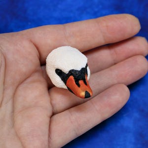 Könnte beinhalten: Eine Miniatur-Schwanenkopf-Figur mit weißem, strukturiertem Körper, schwarzen Augen und einem orange-schwarzen Schnabel. Der Schwan ist ein detailliertes, handgefertigtes Objekt, wahrscheinlich ein Dekorationsobjekt.