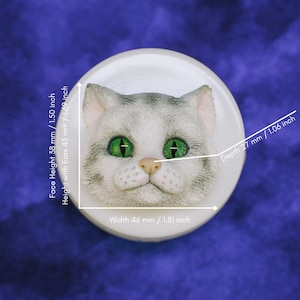 May include: A white, round cat face sculpture with green eyes and a pink nose. The cat's face is detailed with gray and white fur. The sculpture is 1.81 inches wide, 1.50 inches high, and 1.06 inches deep.