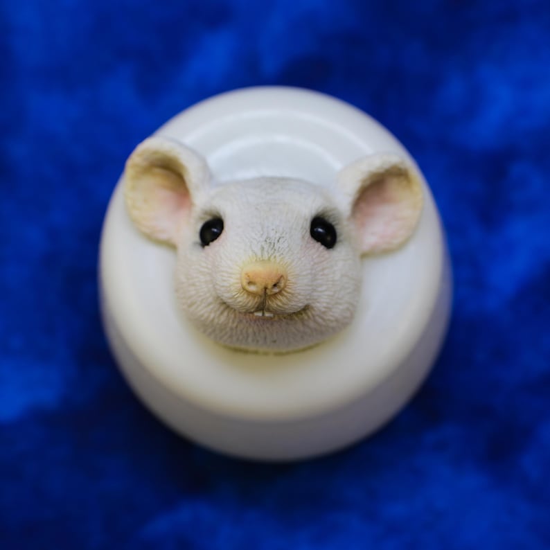 K&ouml;nnte beinhalten: Ein wei&szlig;es, rundes Dekorationsobjekt mit einem detaillierten, l&auml;chelnden Mausgesicht. Die Maus hat rosa Ohren und Nase, schwarze Augen und zwei kleine Z&auml;hne. Das Objekt befindet sich vor einem blauen Hintergrund.