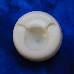 K&ouml;nnte beinhalten: Eine runde, cremefarbene Form mit einem B&auml;rengesicht-Design. Die Merkmale des B&auml;ren sind dezent vertieft, wodurch ein dreidimensionaler Effekt entsteht. Die Form befindet sich vor einem tiefblauen Hintergrund.