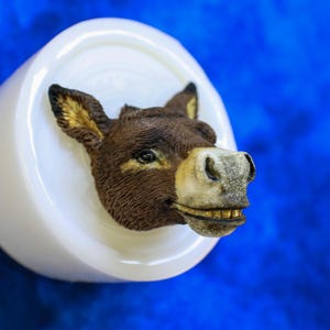 May include: A detailed, brown donkey head sculpture with a smiling expression, set against a white circular base. The donkey's ears have gold accents, and the background is a vibrant blue.