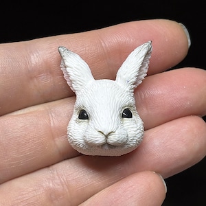 Peut inclure: Figurine de tête de lapin blanche avec des yeux noirs et une texture de fourrure détaillée. Les oreilles du lapin ont des pointes grises. La figurine est tenue dans une main, mettant en valeur la petite taille et le design complexe du lapin.