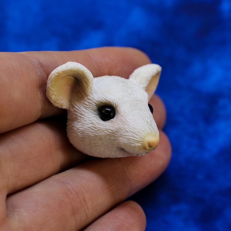 K&ouml;nnte beinhalten: Detaillierte, wei&szlig;e M&auml;usefigur mit schwarzen Augen und einer hellbraunen Nase und Ohren. Das Fell der Maus ist strukturiert, und die Figur wird in einer Hand vor blauem Hintergrund gehalten. Miniatur-Skulptur.
