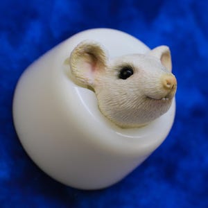 K&ouml;nnte beinhalten: Ein wei&szlig;es, eif&ouml;rmiges Deko-Objekt mit einem detaillierten M&auml;usekopf, der aus der Oberseite herausragt. Die Maus hat rosa Ohren und Nase, schwarze Augen und einen leicht ge&ouml;ffneten Mund. Der Hintergrund ist einfarbig, tiefblau.