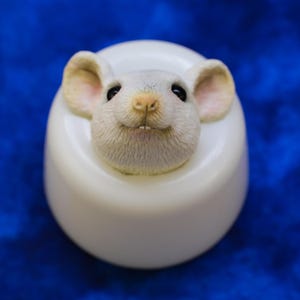 K&ouml;nnte beinhalten: Ein wei&szlig;er Eierbecher aus Keramik mit einem detaillierten, l&auml;chelnden M&auml;usegesicht, das aus der Oberseite herausragt. Die Maus hat rosa Ohren und eine rosa Nase. Der Hintergrund ist tiefblau.