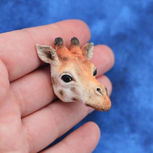 Puede incluir: Una detallada figura en miniatura de la cabeza de una jirafa. La jirafa tiene una combinación de colores marrón y naranja con ojos negros y orejas pequeñas. La figura se sostiene en una mano sobre un fondo azul.
