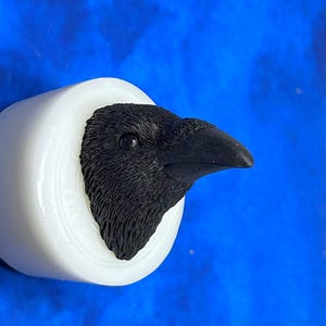 Könnte beinhalten: Eine schwarze Rabenkopfskulptur, die aus einer weißen zylindrischen Form hervortritt. Der Rabenkopf ist mit strukturierten Federn und einem scharfen Schnabel detailliert. Der Hintergrund ist ein leuchtendes Blau.