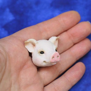Puede incluir: Una pequeña figura de cabeza de cerdo blanca con hocico y orejas rosas. El cerdito tiene ojos negros y una ligera sonrisa. La figura se sostiene en una mano sobre un fondo azul. Es un artículo decorativo en miniatura.
