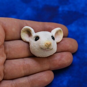 K&ouml;nnte beinhalten: Eine wei&szlig;e M&auml;usefigur mit schwarzen Augen und einer rosa Nase. Die Maus hat gro&szlig;e, abgerundete Ohren und einen l&auml;chelnden Ausdruck. Die Figur wird in einer Hand vor blauem Hintergrund gehalten.