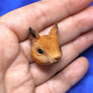 Puede incluir: Una detallada figura en miniatura de una cabeza de ardilla. La ardilla tiene pelaje marrón con detalles negros en las orejas y alrededor de los ojos. La figura se sostiene en una mano, mostrando los intrincados detalles de la escultura.