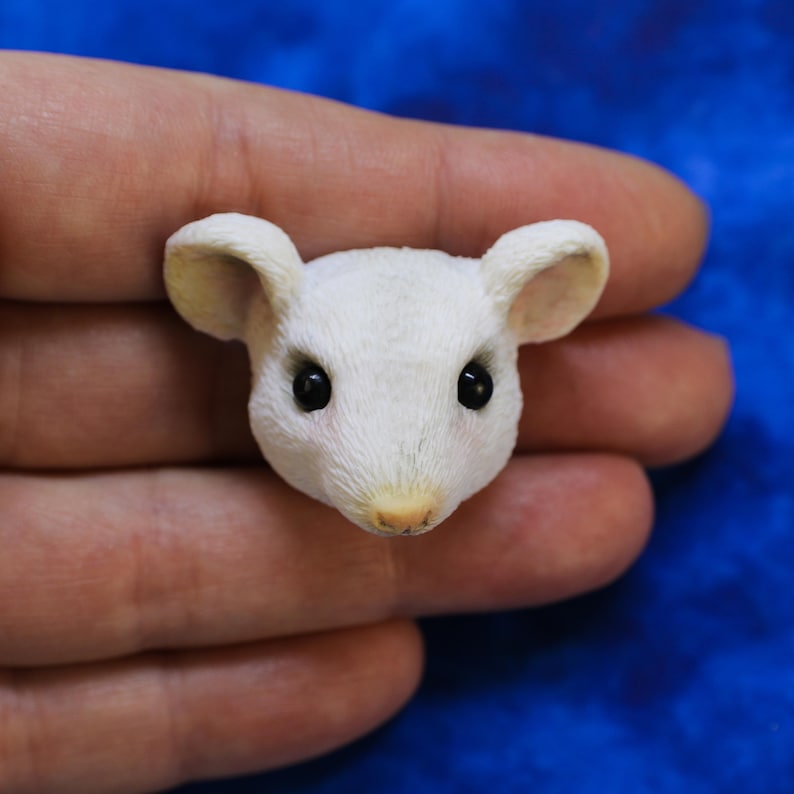 K&ouml;nnte beinhalten: Ein detaillierter, miniaturisierter wei&szlig;er M&auml;usekopf mit schwarzen Augen und einer hellrosa Nase. Die Maus hat gro&szlig;e, abgerundete Ohren und strukturiertes Fell. Die Skulptur wird in einer Hand vor blauem Hintergrund gehalten, was die Details zeigt.