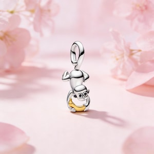 Puede incluir: Un charm de plata con forma de mono sosteniendo un plátano dorado. El mono está en una pose juguetona, con los brazos levantados. El charm tiene un lazo en la parte superior para sujetarlo a una pulsera o collar. El fondo es rosa suave.