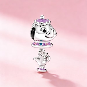 Puede incluir: Un charm de plata con un diseño de tetera y taza. La tetera tiene una tapa y un cuello morados con detalles rosas, azules y morados. La taza cuelga debajo, también plateada con detalles morados. El charm está sobre un fondo rosa.