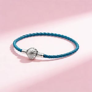 Könnte beinhalten: Ein türkisblaues geflochtenes Lederarmband mit einem silberfarbenen Verschluss. Der Verschluss hat ein kugelförmiges Design mit einem Spiralmuster. Das Armband wird vor einem rosa Hintergrund gezeigt.