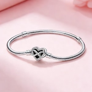 Könnte beinhalten: Ein silbernes Armband mit einem herzförmigen Anhänger. Der Anhänger zeigt ein Unendlichkeitssymbol und ist mit kleinen, klaren Steinen besetzt. Das Armband hat ein Schlangenketten-Design und wird auf einem weichen rosa Hintergrund präsentiert.