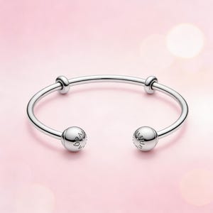 May include: A silver open bangle bracelet with two spherical end caps and two smaller spherical accents. The bracelet has a smooth, polished finish against a soft pink background.