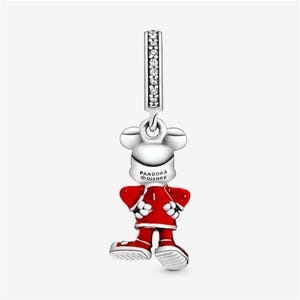 Puede incluir: Un charm de plata con una figura estilizada de Mickey Mouse con un atuendo rojo. El charm tiene un dise&ntilde;o colgante con una barra vertical adornada con piedras transparentes. La parte posterior del charm tiene el texto "PANDORA Disney".