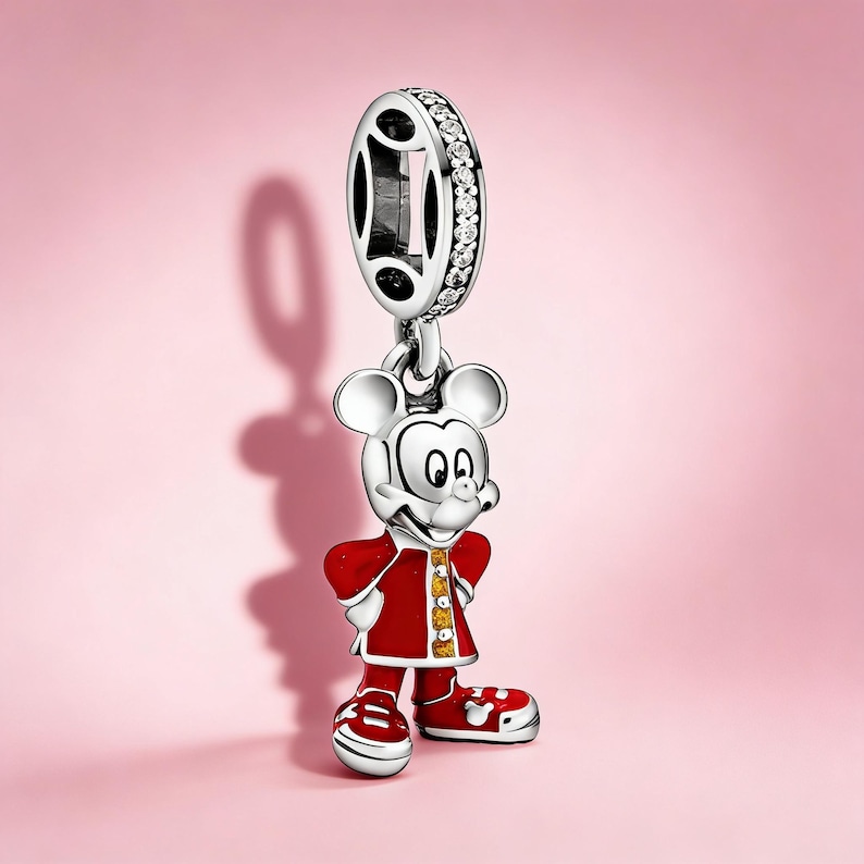 Puede incluir: Un charm de plata con Mickey Mouse con abrigo y zapatos rojos con detalles blancos. El charm tiene una parte superior decorativa con piedras transparentes y detalles negros. El fondo es rosa liso.