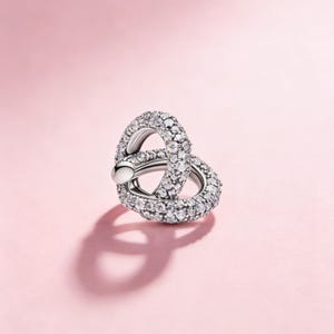 Op de afbeelding: Een zilverkleurige, verweven ring, bezet met talrijke kleine, heldere edelstenen. De ring heeft een gladde, gepolijste afwerking en staat tegen een zachtroze achtergrond. Het ontwerp heeft twee in elkaar grijpende lussen, die een unieke vorm creëren.