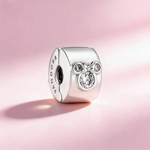May include: A silver charm with a bow design and three clear, sparkling stones. The charm is cylindrical with the word "PANDORA" engraved on the side. The charm is likely made of sterling silver.