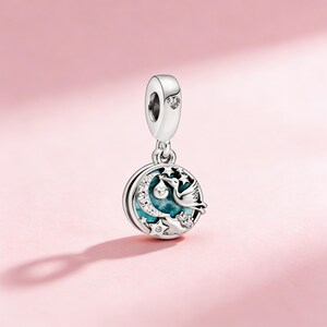 May include: A silver charm with a stork carrying a baby, set against a crescent moon and stars. The charm has a turquoise enamel background and is accented with clear crystals. The top loop features a heart-shaped crystal.