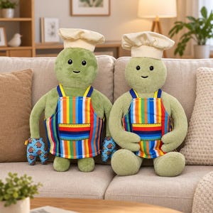 Pode incluir: Duas figuras de peluche verdes a usar chapéus de chef cor de creme e aventais às riscas verticais coloridas. As figuras têm traços faciais bordados simples e luvas de forno azuis com um padrão de aves kiwi castanhas nas mãos.