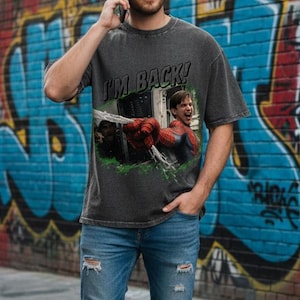 May include: A dark gray t-shirt with the text "I'M BACK!" and a graphic of a superhero. The person is wearing blue jeans with distressed details and white sneakers. The background features a brick wall covered in colorful graffiti.