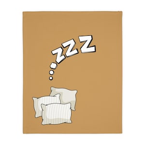 May include: A tan blanket featuring a cartoon illustration of three pillows and a thought bubble with the word "ZZZ" in white letters. The pillows are light beige with white stripes.