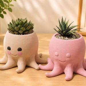 Può includere: Due vasi per piante a forma di polpo in materiale opaco. Il vaso a sinistra è beige con occhi neri e guance rosa, contenente una succulenta verde. Il vaso a destra è rosa con occhi chiusi e bocca sorridente, contenente una succulenta verde.
