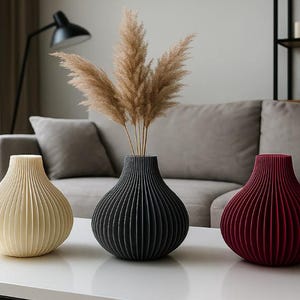 May include: Three decorative vases in cream, black, and burgundy, each with a ribbed design. The vases are arranged on a white table, with dried ornamental grass in the black vase. A modern home decor theme.