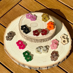 Peut inclure: Une collection de bagues en forme de fleurs de différentes couleurs, dont rose, or, orange et vert, présentées sur une tranche de bois. Les bagues sont faites de différents matériaux, comme des paillettes et du bois. Le fond est une surface en bois.