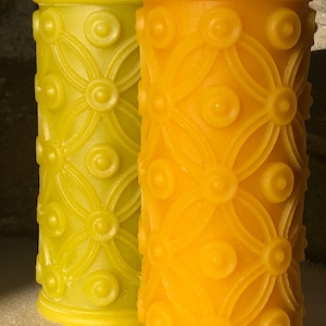 May include: Two decorative pillar candles with a geometric pattern. The candle on the left is green and the candle on the right is yellow.