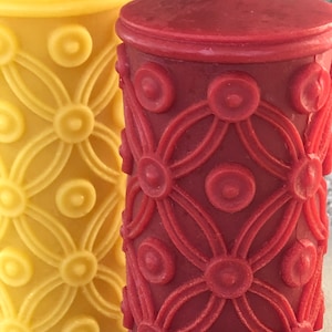 May include: Two pillar candles with a decorative floral pattern. The candle on the left is yellow and the candle on the right is red.