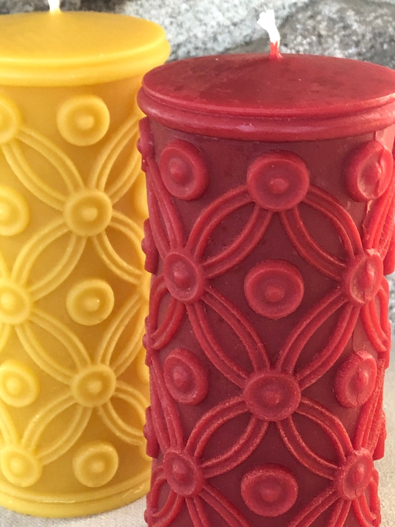 May include: Two pillar candles with a decorative floral pattern. The candle on the left is yellow and the candle on the right is red.