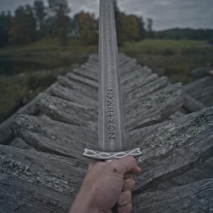 May include: A silver sword with the word "NORTHMEN" etched on the blade is held up against a cloudy sky. The sword has a decorative handle and is held over a weathered wooden structure.