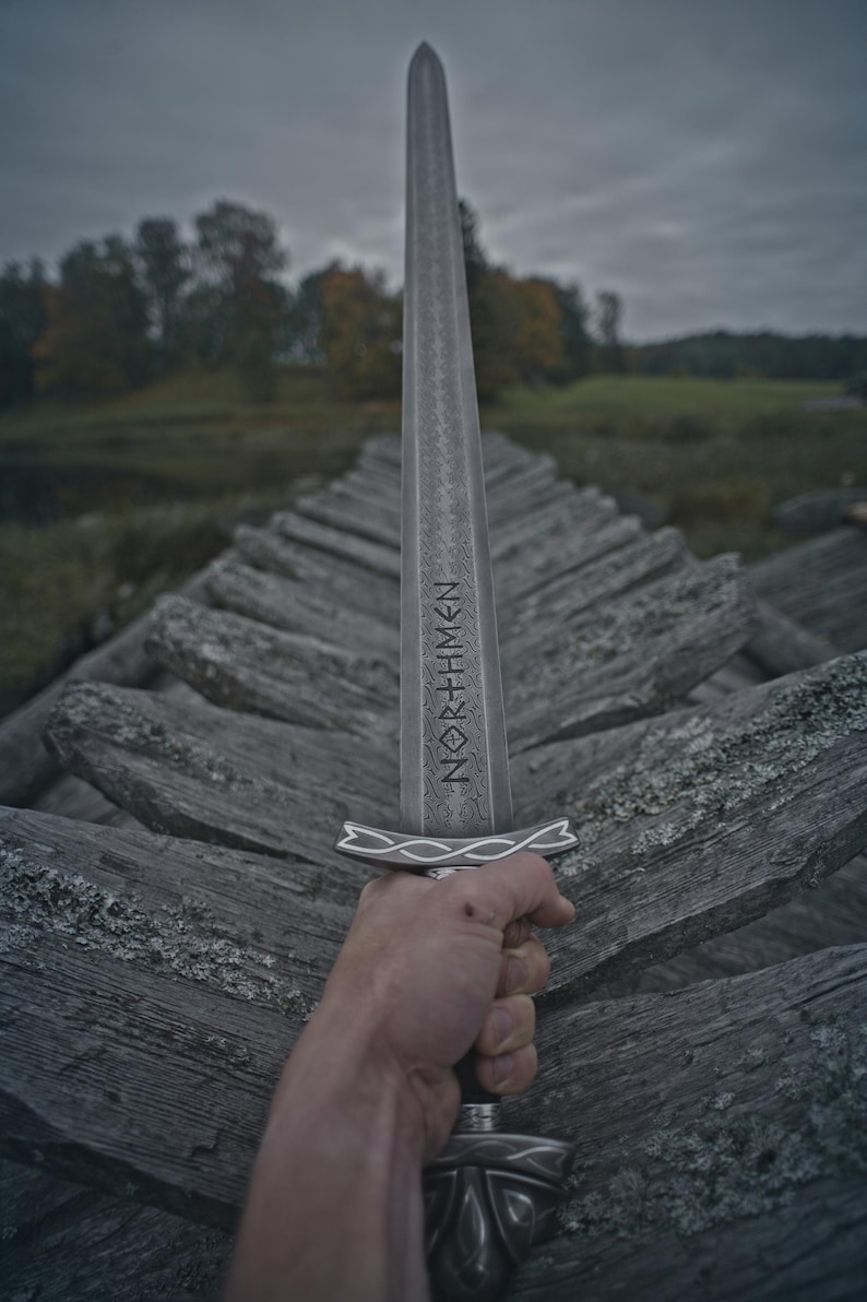 May include: A silver sword with the word "NORTHMEN" etched on the blade is held up against a cloudy sky. The sword has a decorative handle and is held over a weathered wooden structure.