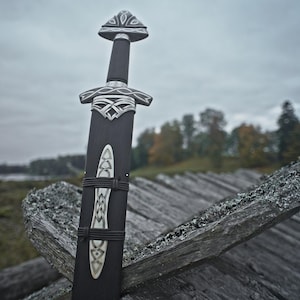 May include: A black sword with ornate silver detailing, resting against a weathered wooden structure. The sword's handle and sheath are dark, with intricate patterns. The background features a cloudy sky and a landscape with trees.
