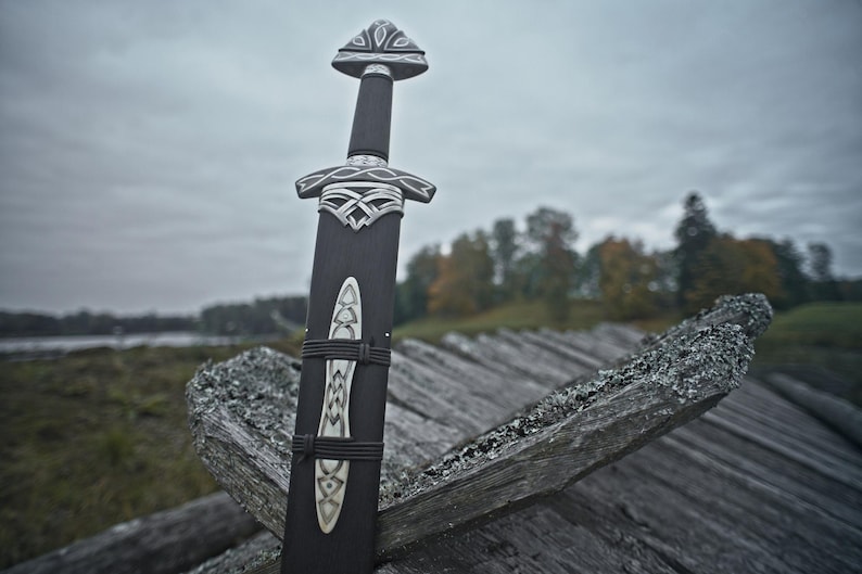 May include: A black sword with ornate silver detailing, resting against a weathered wooden structure. The sword's handle and sheath are dark, with intricate patterns. The background features a cloudy sky and a landscape with trees.