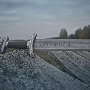 May include: A detailed image of a Viking sword with the word "NORTHMEN" etched into the blade. The sword rests on a weathered wooden surface, with a dark handle and ornate silver-colored hilt. The background features a blurred landscape under a cloudy sky.