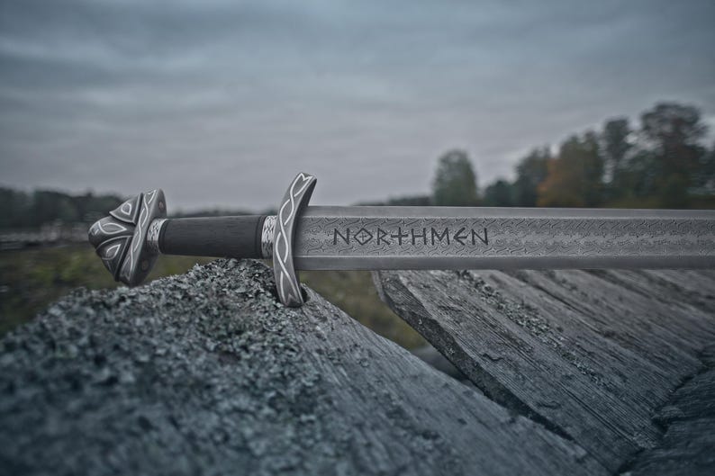 May include: A detailed image of a Viking sword with the word "NORTHMEN" etched into the blade. The sword rests on a weathered wooden surface, with a dark handle and ornate silver-colored hilt. The background features a blurred landscape under a cloudy sky.