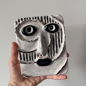 May include: A ceramic face sculpture with a rectangular shape. The sculpture features black and white colors, with black detailing for the eyes, mouth, and vertical lines. The face has a textured surface and a unique, artistic design.