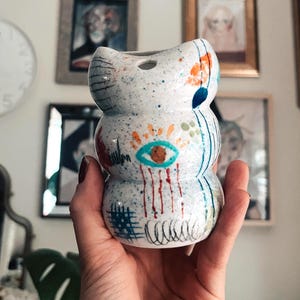 Peut inclure: Petit vase en céramique blanche avec une finition mouchetée et des détails peints abstraits. Le vase présente un design empilé avec une large ouverture et est orné d'accents bleus, orange et rouges. Le vase est tenu dans une main.