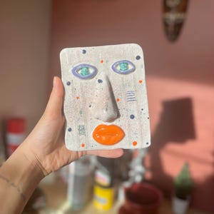 Op de afbeelding: Een rechthoekige keramische gezichtssculptuur met een getextureerd, gebroken wit oppervlak. Het gezicht heeft grote, turquoise ogen met paarse omtreklijnen, een oranje mond en een prominente neus. De sculptuur is versierd met blauwe, oranje en groene stippen.